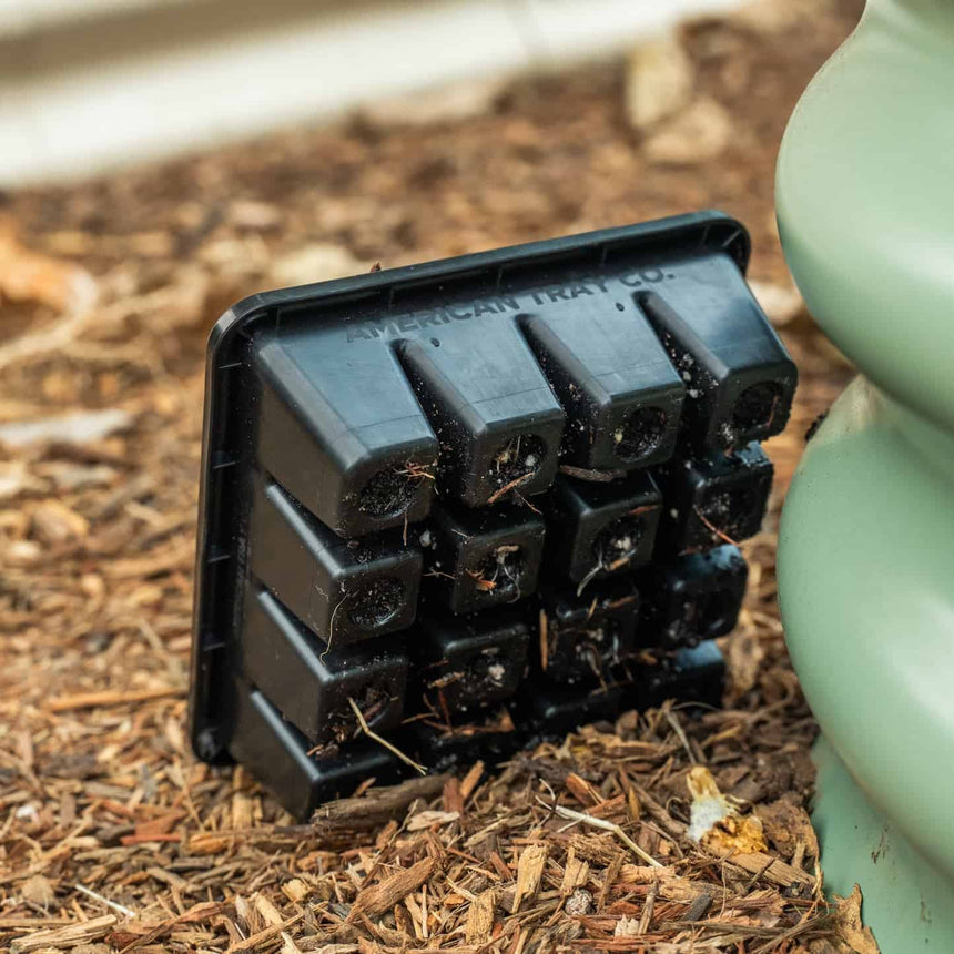 An empty black 16-cell seed starting tray, embossed with the American Tray Co. logo and flecked with dried soil, leaning against a pale green planter on a bed of wood mulch.