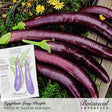 Several glossy, dark purple Long Purple eggplants laid out on a dark wooden surface with green leaves, yellow flowers, and a Botanical Interests seed packet, labeled "Eggplant Long Purple / Photo by Sujatha Marsden" and "Botanical Interests" for Long Purple Eggplant Seeds.