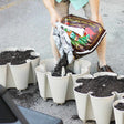 A person pours dark potting soil from a labelled garden soil bag into the scalloped, multi-pocket tiers of a stacked cream-coloured GreenStalk vertical planter set on pavement.