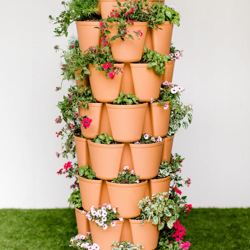 Terracotta-coloured GreenStalk 7-tier vertical planter tower, each staggered pocket planted with a mix of trailing greenery and flowering plants including pink verbena and white petunias, standing on artificial grass against a plain wall.