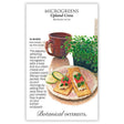 Seed packet illustration for Upland Cress Microgreens showing a tray of leafy green sprouts beside a mug, with crackers topped with cream cheese, tomato, and cress on a floral plate; text notes a 14–28 day growing time and describes the peppery flavor with a suggested recipe.