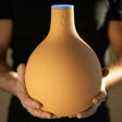 A person holds a bulbous, unglazed terracotta Garden Oya watering pot with a blue rubber cap and a small teardrop symbol etched near its narrow neck.