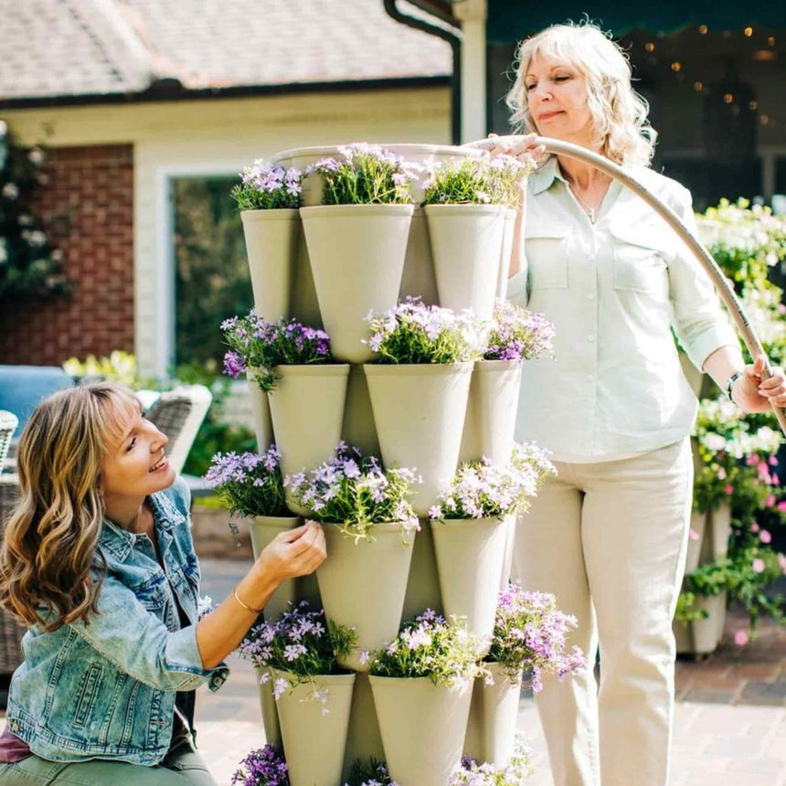Two women tend a stacked GreenStalk 5-Tier Vertical Planter in cream, its five layers of angled pockets overflowing with trailing purple phlox, one woman watering with a hose while the other adjusts blooms on a sunny patio.