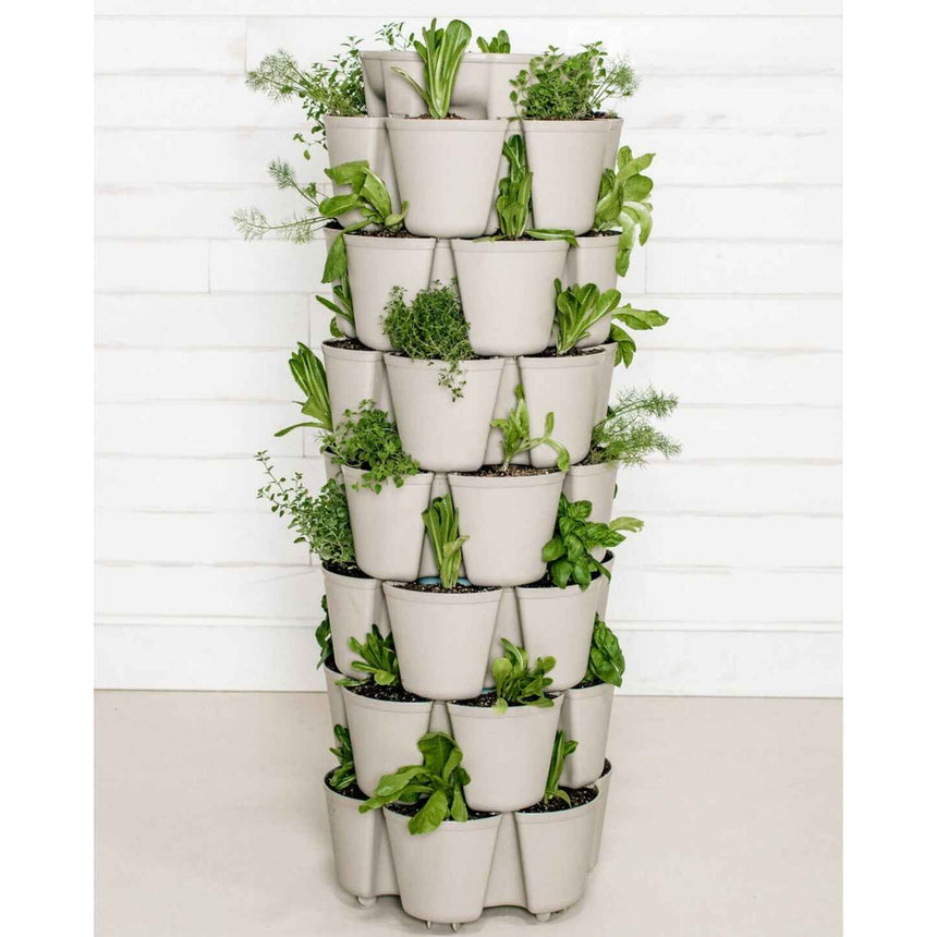 Tall stackable vertical planter with six tiers of overlapping stone-grey pockets on a mobile base, densely planted with leafy herbs and greens like thyme, basil and lettuce, set against a white shiplap wall.