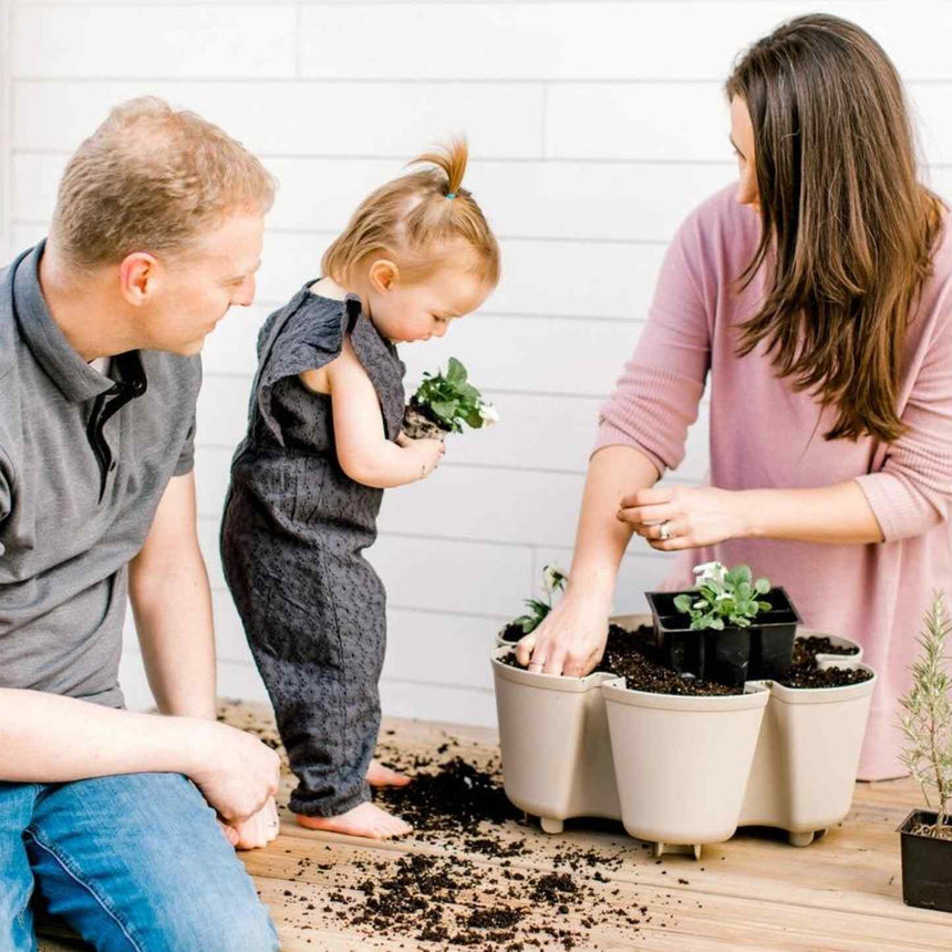 A toddler in overalls holds a small flowering plant while her parents kneel beside her on a wooden deck, planting seedlings into the scalloped, clover-shaped pots of a cream multi-pocket planter, with soil scattered around.
