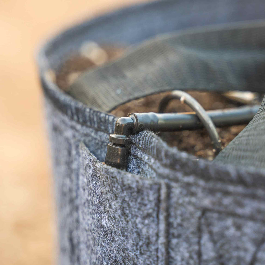 Close-up of a grey felt grow bag filled with soil, showing a drip irrigation elbow fitting and tubing threaded through the fabric rim.