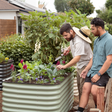 Two men, one wearing a straw hat and holding a trowel, tend kale, snapdragons, and pansies growing in a tall sage-green corrugated metal raised garden bed in a lush backyard garden.