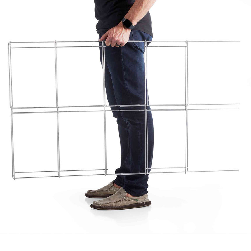 A person in jeans holds a folded wire tomato cage upright against their leg to demonstrate its full extended height and grid-panel construction.