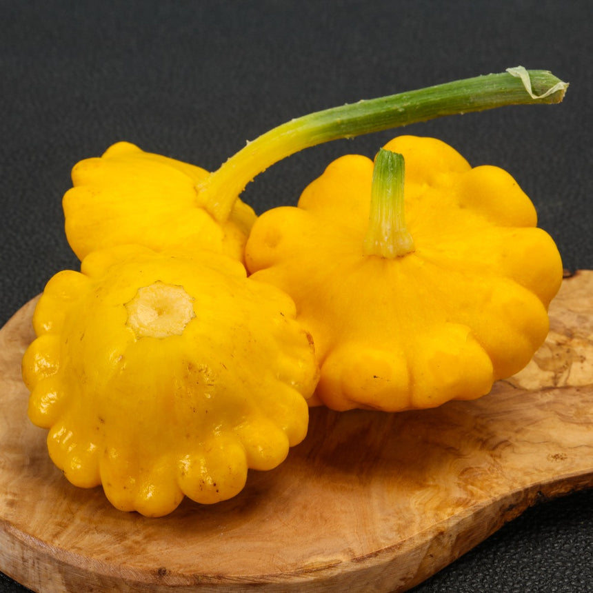 Three bright yellow, scalloped Gelber Englischer pattypan squashes resting on a rustic wooden cutting board against a dark background.