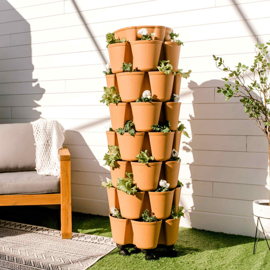 A tall terracotta-colored GreenStalk 7-tier vertical planter on wheels, its stacked pockets filled with leafy greens and white flowers, standing beside an outdoor sofa on artificial turf against a white shiplap wall.