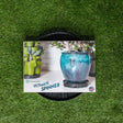 A round black plastic GreenStalk Ultimate Spinner base sits on artificial turf, its packaging showing a glazed blue-grey planter and a stacked green vertical garden with vegetables growing.