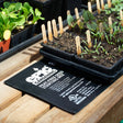 Black seed trays of young seedlings labelled with wooden plant markers sit atop an Epic Gardening heat mat printed with a peat-free seed starting mix recipe and appliance specifications, on a wooden potting bench.