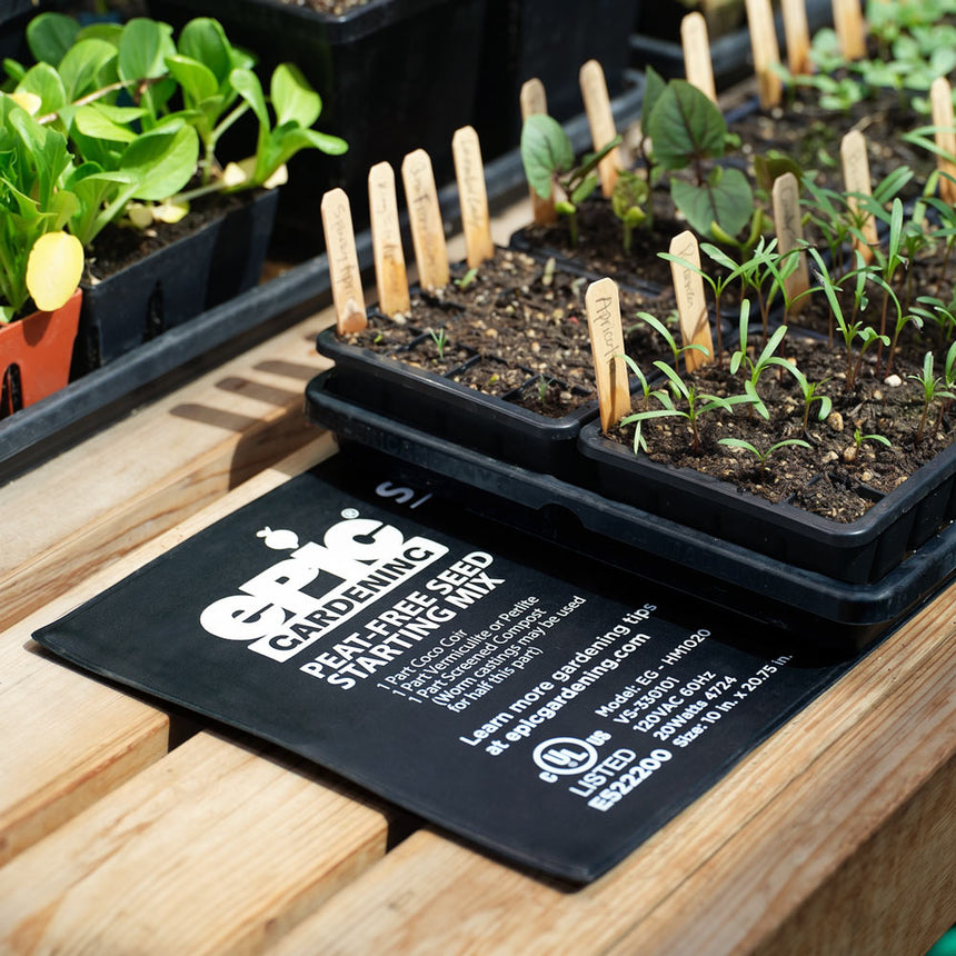 Black seed trays of young seedlings labelled with wooden plant markers sit atop an Epic Gardening heat mat printed with a peat-free seed starting mix recipe and appliance specifications, on a wooden potting bench.
