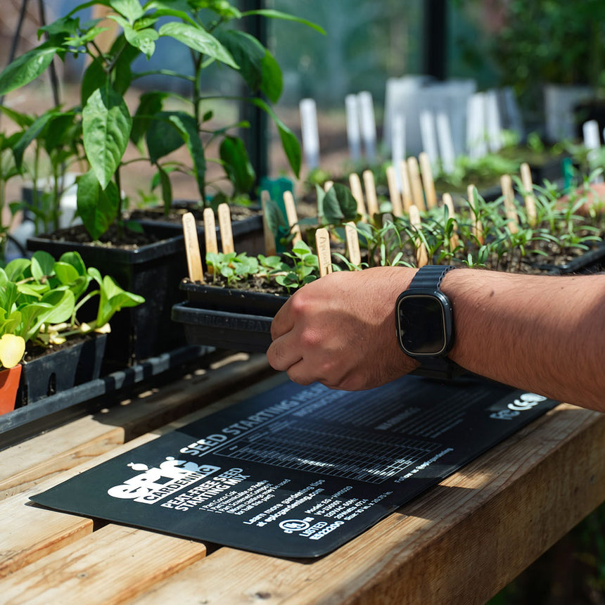A person's wrist rests near a black Epic Gardening seed starting heat mat printed with usage instructions and a temperature chart, resting on a wooden greenhouse bench beside trays of labelled seedlings.