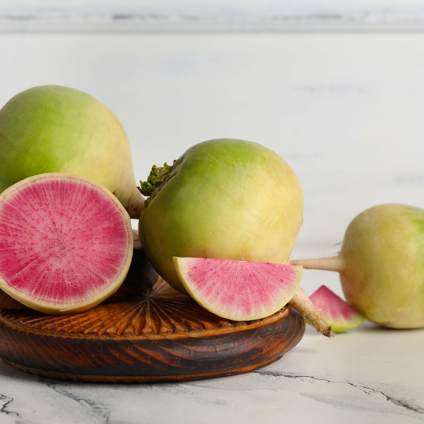 Whole watermelon radishes with pale green-and-white skin arranged on a wooden dish, with one halved and one wedge cut open to reveal vivid pink, radiating flesh inside.