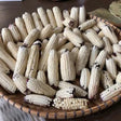 Woven basket piled high with small husked corn cobs showing pearly white kernels, some tipped with dark purple-black coloring near the base.