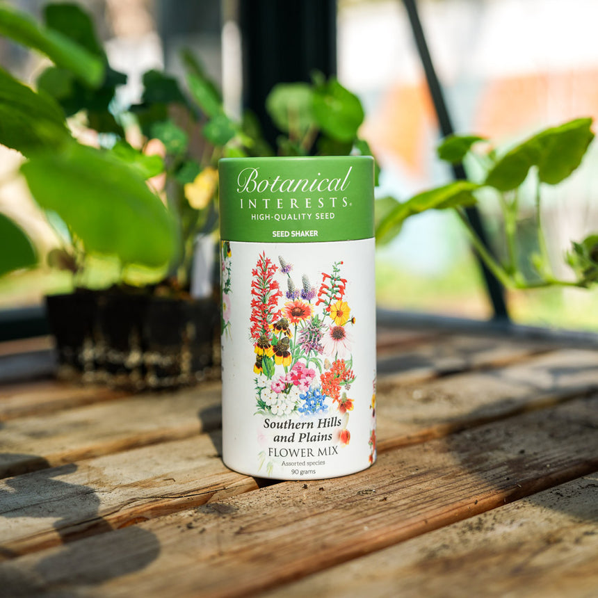 Botanical Interests Southern Hills and Plains Flower Mix Seed Shaker canister on a wooden surface with plants in the background