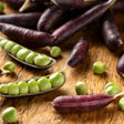 Purple Sugar Magnolia snap pea pods scattered on a wooden board, some split open to reveal rows of plump bright green peas inside.