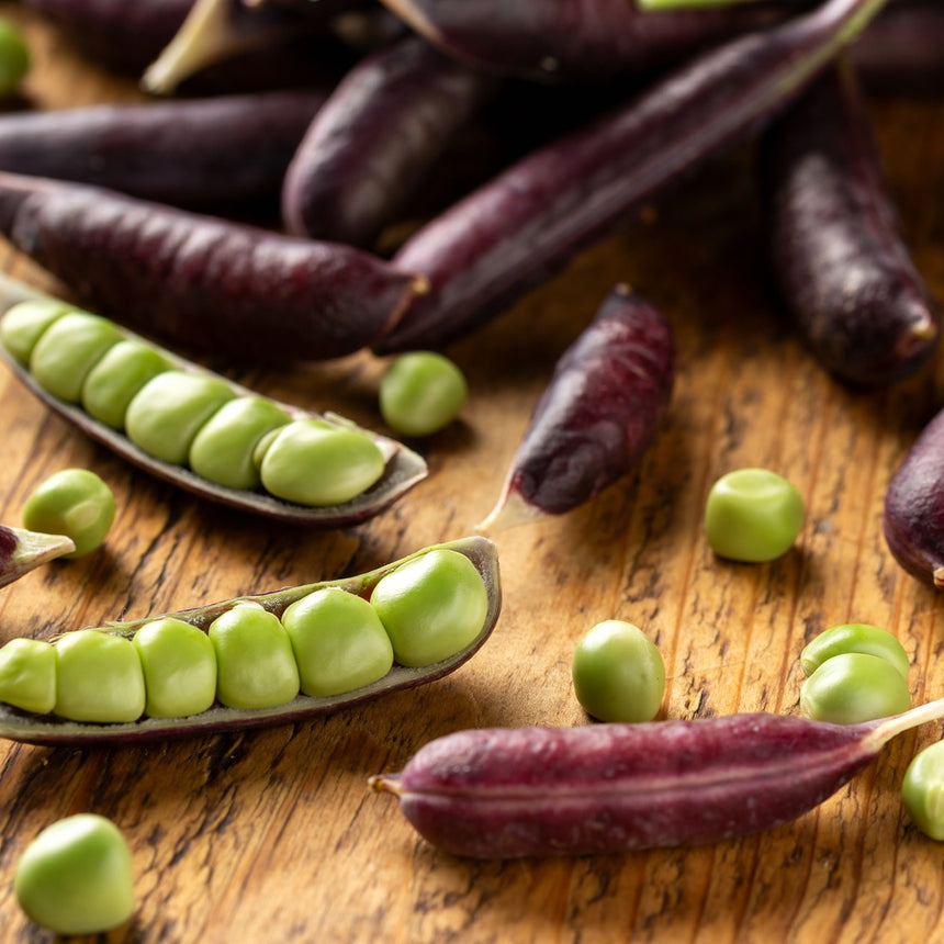 Purple Sugar Magnolia snap pea pods scattered on a wooden board, some split open to reveal rows of plump bright green peas inside.
