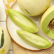 Whole and sliced honeydew melons with pale yellow-green skin and light green flesh, some seeded wedges peeled and others showing seed cavities, arranged on a plate atop a yellow-checked cloth.